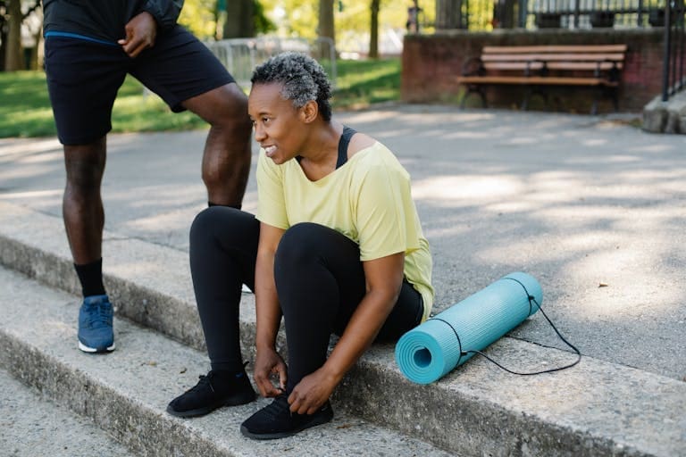 A mature African American woman ties shoelaces beside a yoga mat outdoors.